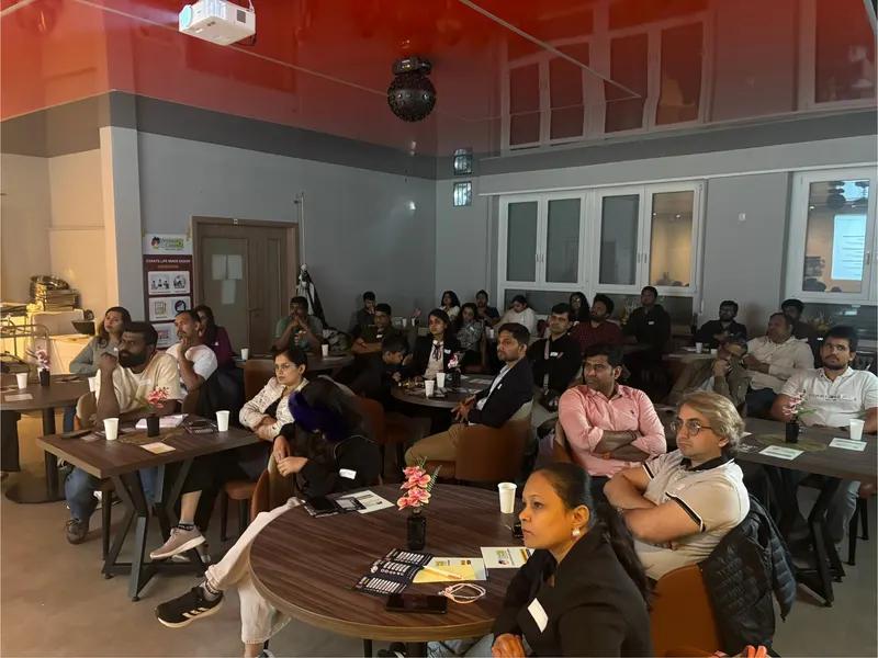 Attendees seated at multiple round tables with name badges and refreshments listen during a presentation.