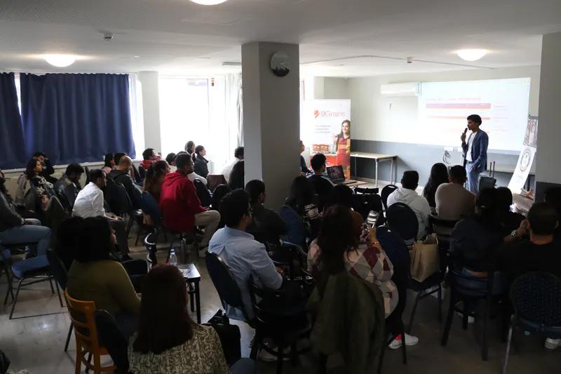 A speaker presents to a full room of seated attendees in a bright seminar room, with a slide screen and roll-up banner at the front.